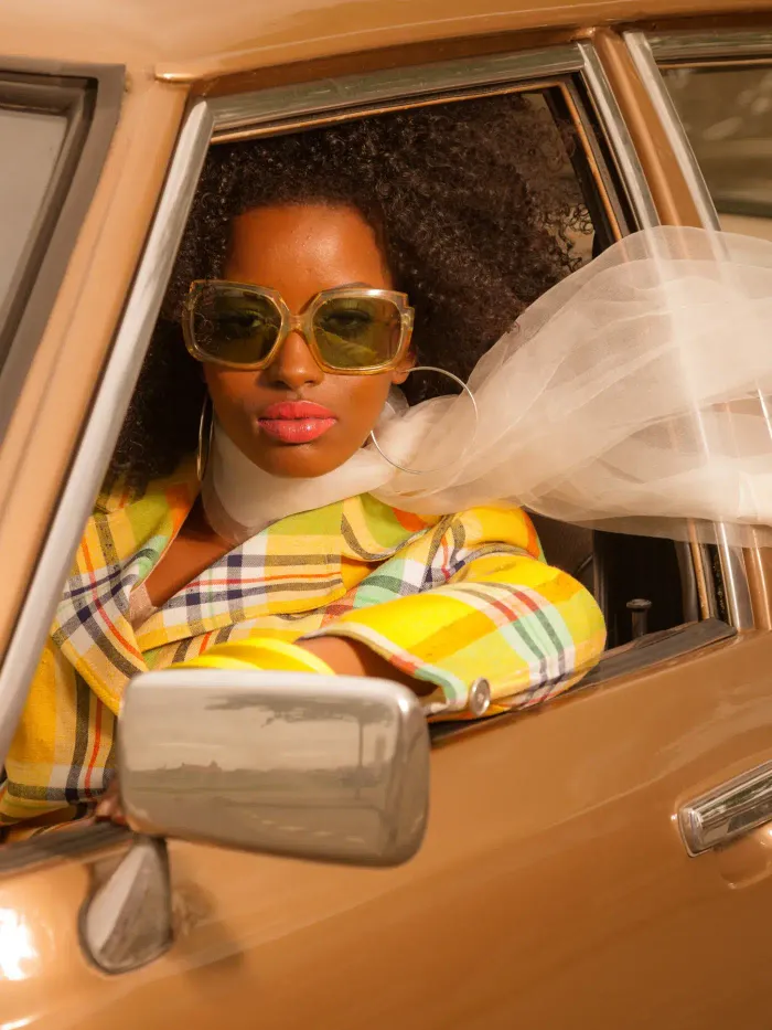 Femme avec de grosses lunettes de soleil, du rouge à lèvre rose, des grandes créoles et un foulard qui vole par la fenêtre ouverte au volant d'une voiture des années 70